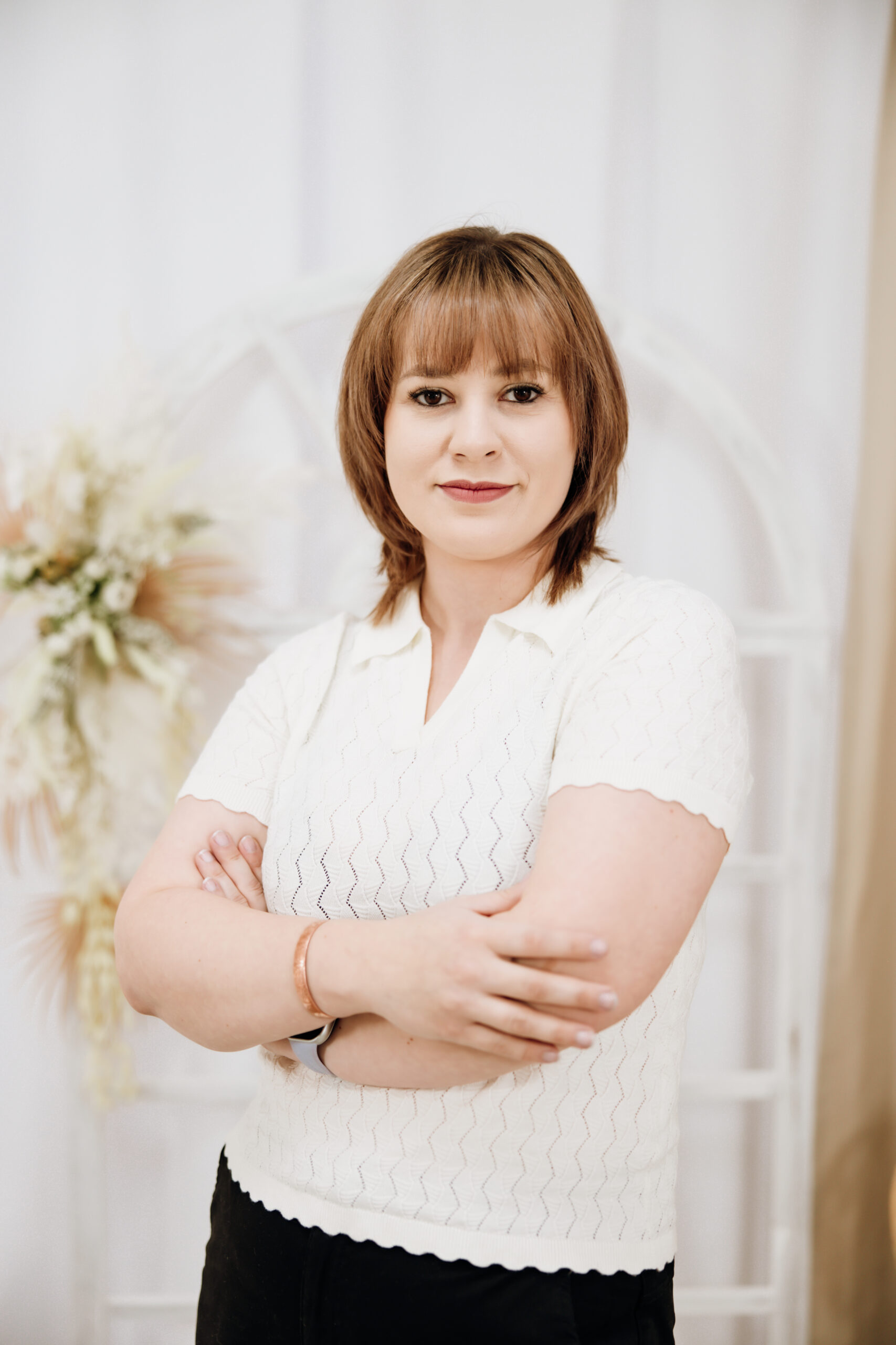 Kobieta z brązowymi włosami i grzywką w białej bluzce z krótkim rękawem, stojąca z założonymi rękami na jasnym, rozmytym tle.