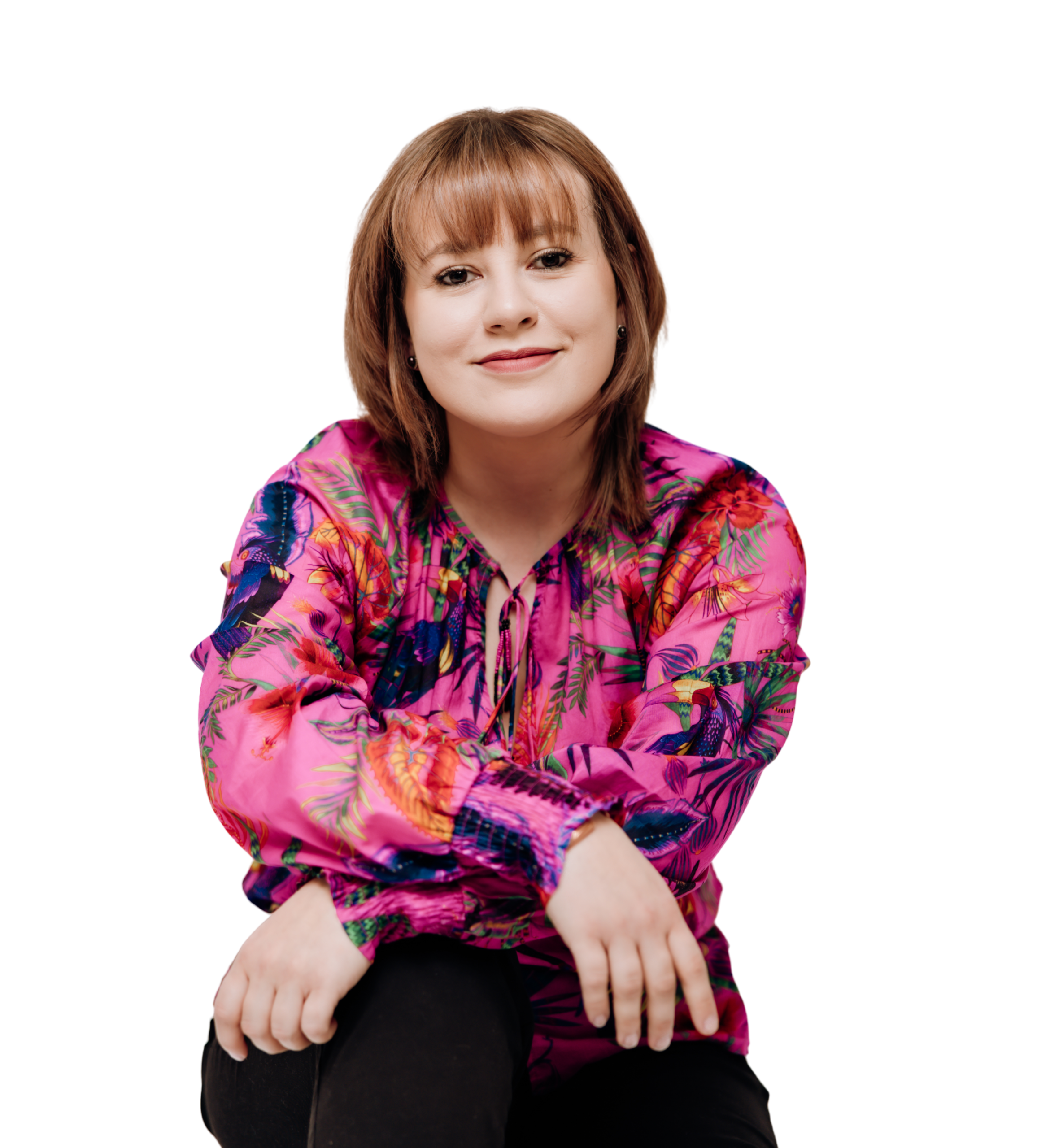 Kobieta o krótkich brązowych włosach z grzywką, ubrana w różową koszulę w kolorowe, roślinne wzory, siedząca na białym tle.